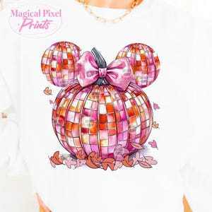 K&ouml;nnte beinhalten: Ein rosa und orangefarbener K&uuml;rbis mit einer rosa Schleife, die wie Mickey-Maus-Ohren geformt ist, mit Herbstbl&auml;ttern, die um ihn herum verstreut sind.