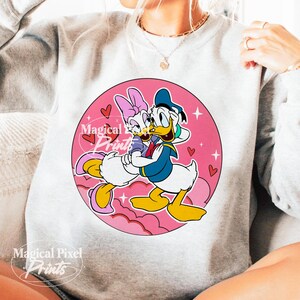 以下が含まれることがあります： ピンクの円形のグラフィックが施された白いスウェットシャツ。グラフィックには、ロマンチックな抱擁をするドナルドダックとデイジーダックが描かれています。グラフィックには、ピンクのハートと星が描かれています。画像の下部には「Magical Pixel Prints」というテキストが表示されています。