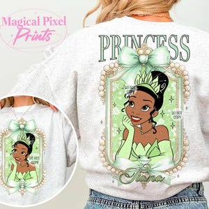 May include: A gray sweatshirt with a green princess design. The design features a portrait of a cartoon princess with a green dress and crown, surrounded by a gold frame with pearls. The text "PRINCESS" is printed in green above the design, and the name "Tiana" is printed below the design.