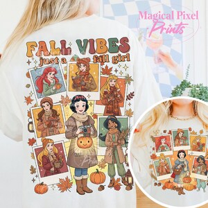 May include: Cream-colored t-shirt with a graphic featuring cartoon princesses in fall attire. The shirt has the text "FALL VIBES just a fall girl" in orange and brown. The design includes pumpkins and autumn leaves.