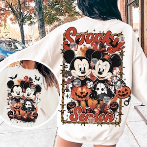 May include: Cream-colored sweatshirt featuring a Halloween-themed graphic. The design includes Mickey and Minnie Mouse dressed in Halloween costumes, surrounded by pumpkins and the words "Spooky Season" in orange and red. The image is framed by a barbed wire design.