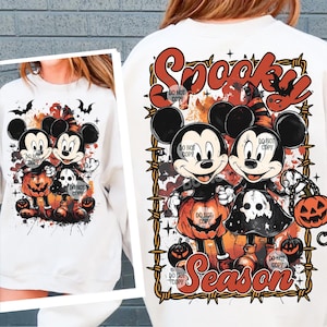 May include: White sweatshirt featuring a Halloween-themed design with Mickey and Minnie Mouse characters dressed in costumes, holding jack-o'-lanterns. The words "Spooky Season" are displayed in a decorative font, with bats and barbed wire accents.