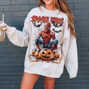 K&ouml;nnte beinhalten: Cremefarbenes Sweatshirt mit einer Grafik von Spider-Man auf K&uuml;rbissen, dem Text "Spooky Vibes" und Flederm&auml;usen. Das Design hat einen Vintage-Comic-Stil.
