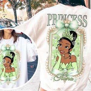 May include: White sweatshirt with a green and gold princess design featuring a cartoon image of Tiana from Disney's The Princess and the Frog. The text "Princess" is printed above the design and "Tiana" is printed below the design.
