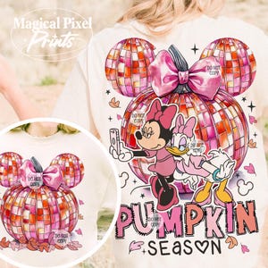 K&ouml;nnte beinhalten: Ein wei&szlig;es T-Shirt mit einem pinkfarbenen und orangenen K&uuml;rbisdesign. Der K&uuml;rbis ist in Form von Mickey-Mouse-Ohren und hat ein Discokugelmuster. Mickey Mouse und Daisy Duck stehen auf dem K&uuml;rbis und machen ein Selfie. Der Text "PUMPKIN SEASON" steht unter dem K&uuml;rbis.