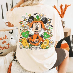 May include: Cream-colored t-shirt featuring a Halloween-themed Minnie Mouse design. Minnie is dressed as a pumpkin, surrounded by flowers and the text "Minnie Mouse xoxo". The shirt has the text "Do Not Copy" repeated around the design.