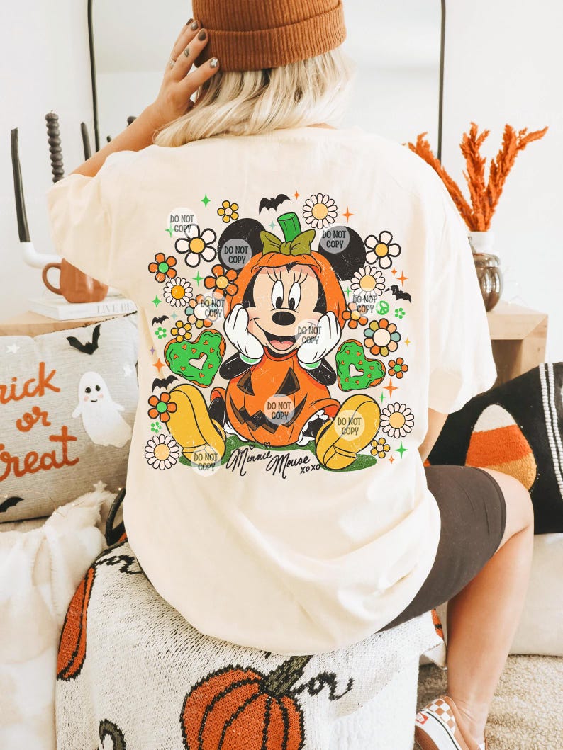 May include: Cream-colored t-shirt featuring a Halloween-themed Minnie Mouse design. Minnie is dressed as a pumpkin, surrounded by flowers and the text "Minnie Mouse xoxo". The shirt has the text "Do Not Copy" repeated around the design.