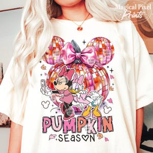 K&ouml;nnte beinhalten: Ein wei&szlig;es T-Shirt mit einem pinkfarbenen und orangenen K&uuml;rbisdesign. Der K&uuml;rbis ist in Form von Mickey-Mouse-Ohren und hat eine rosafarbene Schleife. Zwei Zeichentrickfiguren, Mickey Mouse und Daisy Duck, stehen auf dem K&uuml;rbis. Der Text "PUMPKIN SEASON" ist unter dem K&uuml;rbis geschrieben.