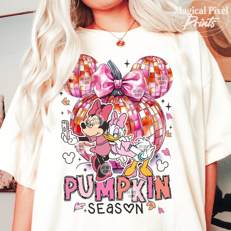 K&ouml;nnte beinhalten: Ein wei&szlig;es T-Shirt mit einem pinkfarbenen und orangenen K&uuml;rbisdesign. Der K&uuml;rbis ist in Form von Mickey-Mouse-Ohren und hat eine rosafarbene Schleife. Zwei Zeichentrickfiguren, Mickey Mouse und Daisy Duck, stehen auf dem K&uuml;rbis. Der Text "PUMPKIN SEASON" ist unter dem K&uuml;rbis geschrieben.