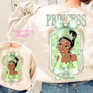 May include: A cream-colored sweatshirt with a green bow and a framed image of Princess Tiana from Disney's The Princess and the Frog. The text "Princess" is printed above the image and "Tiana" is printed below the image.