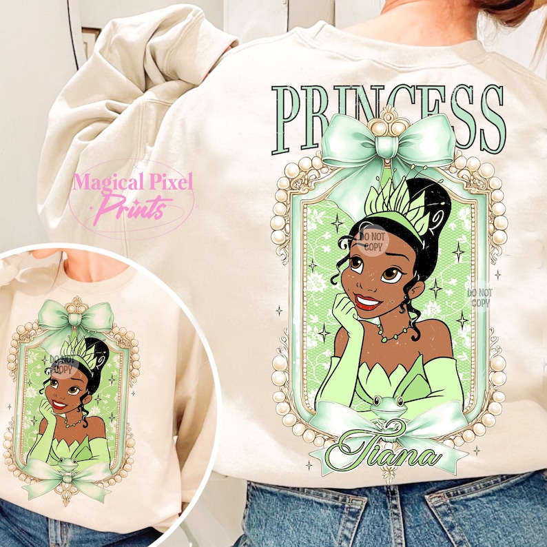 May include: A cream-colored sweatshirt with a green bow and a framed image of Princess Tiana from Disney's The Princess and the Frog. The text "Princess" is printed above the image and "Tiana" is printed below the image.