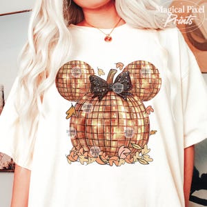 K&ouml;nnte beinhalten: Ein wei&szlig;es T-Shirt mit einem goldenen Glitzerk&uuml;rbis-Design. Der K&uuml;rbis ist in Form von Mickey Mouse Ohren und hat eine schwarze Schleife mit Glitzer. Der K&uuml;rbis ist von Herbstbl&auml;ttern umgeben.