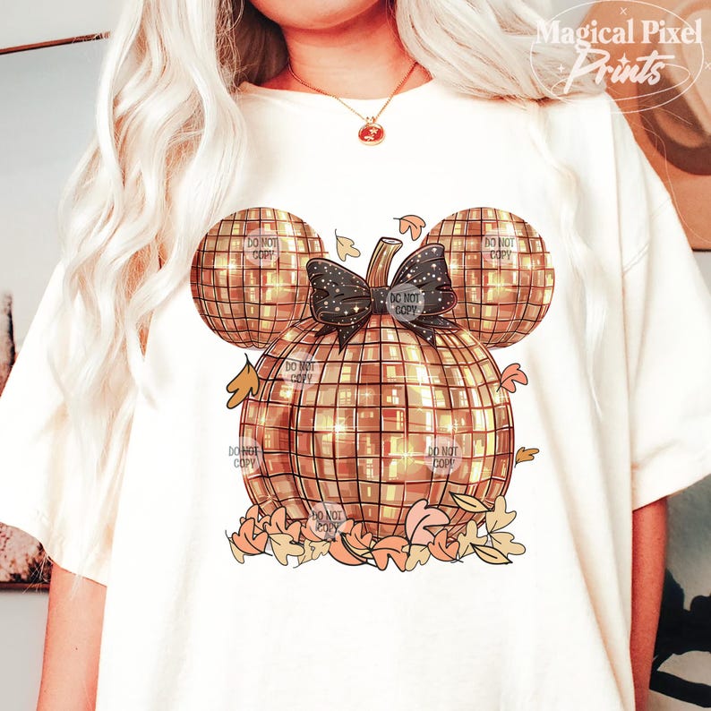 K&ouml;nnte beinhalten: Ein wei&szlig;es T-Shirt mit einem goldenen Glitzerk&uuml;rbis-Design. Der K&uuml;rbis ist in Form von Mickey Mouse Ohren und hat eine schwarze Schleife mit Glitzer. Der K&uuml;rbis ist von Herbstbl&auml;ttern umgeben.