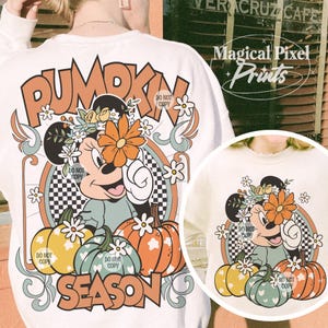 May include: A white sweatshirt with a graphic design featuring a cartoon mouse wearing a flower crown and holding a pumpkin. The text "Pumpkin Season" is printed in orange and white letters. The design is in a retro style with checkered patterns and floral accents.