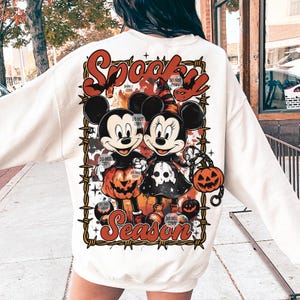 May include: Cream-colored sweatshirt featuring a Halloween-themed graphic. The design includes Mickey and Minnie Mouse dressed in Halloween costumes, surrounded by barbed wire and the words "Spooky Season" in orange and red.