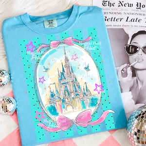 May include: A pale blue t-shirt with a fairytale castle illustration in an oval frame, adorned with pink bows, stars, and birds. The design includes the text "Magical Pixel Prints". The shirt is presented on a pink and white striped surface.