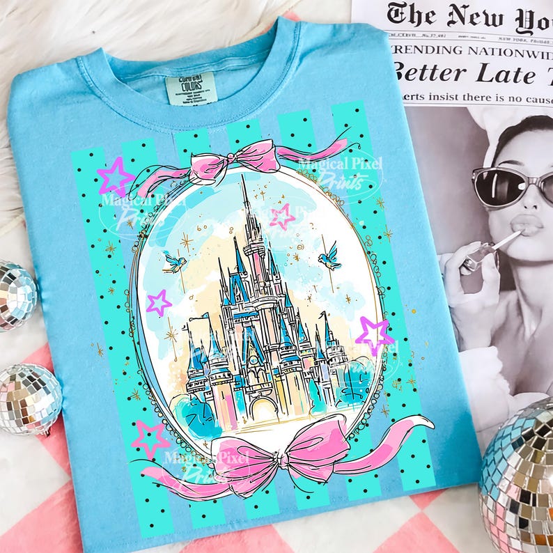 May include: A pale blue t-shirt with a fairytale castle illustration in an oval frame, adorned with pink bows, stars, and birds. The design includes the text "Magical Pixel Prints". The shirt is presented on a pink and white striped surface.