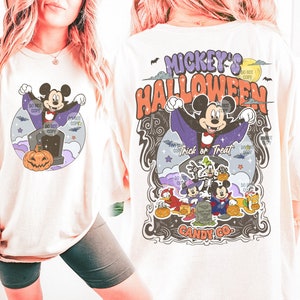 Może przedstawiać: Kremowa koszulka z grafiką o tematyce Halloween. Projekt zawiera Myszka Miki przebranego za wampira, dynię oraz napis "Mickey's Halloween Trick or Treat". Tył koszulki ma podobny wz&oacute;r z innymi postaciami Disneya.