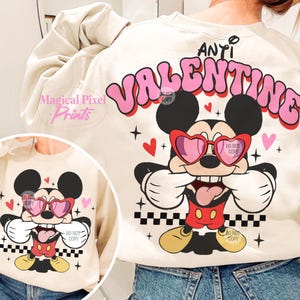 Pu&ograve; includere: Una felpa beige con una grafica rosa che dice "ANTI VALENTINE" con un cartone animato di Topolino che indossa occhiali a forma di cuore e una lingua rossa che sporge. La grafica di Topolino &egrave; su uno sfondo a scacchi con cuori rosa.