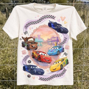 Puede incluir: Camiseta de color crema con un gráfico colorido de coches de carreras animados. El diseño incluye el texto "Route 66" y una bandera a cuadros. Los coches están en un marco circular con un fondo de puesta de sol.