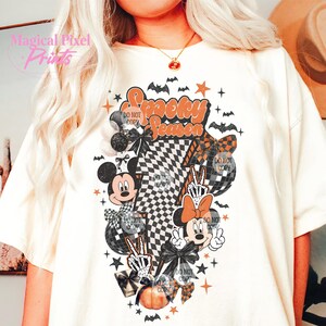 K&ouml;nnte beinhalten: Cremefarbenes T-Shirt mit einem Halloween-Motiv. Das Design umfasst Micky und Minnie Maus, Flederm&auml;use, Sterne, K&uuml;rbisse und den Text "Spooky Season". Die Grafik hat ein Schachbrettmuster und verschiedene dekorative Elemente.
