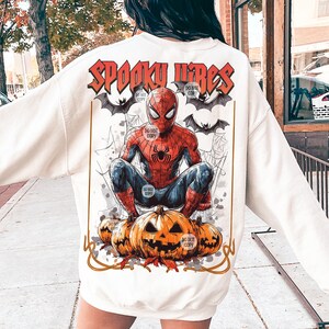 K&ouml;nnte beinhalten: Cremefarbenes Sweatshirt mit einer Grafik von Spider-Man auf K&uuml;rbissen. Das Design umfasst Flederm&auml;use, Spinnweben und den Text "SPOOKY VIBES" in Orange. Ein Kleidungsst&uuml;ck zum Thema Halloween.