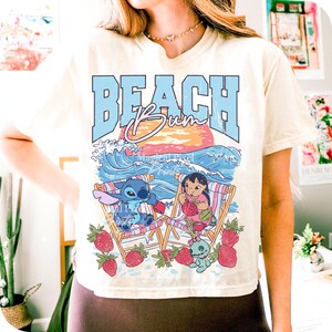Puede incluir: Camiseta color crema con un gr&aacute;fico de Stitch y Lilo en sillas de playa, rodeados de fresas. El texto "BEACH BUM" est&aacute; en azul, sobre un atardecer y olas del oc&eacute;ano. Un dise&ntilde;o divertido y veraniego.