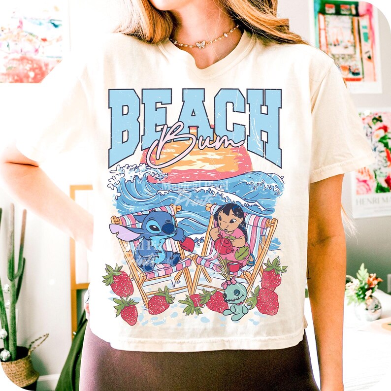 Puede incluir: Camiseta color crema con un gr&aacute;fico de Stitch y Lilo en sillas de playa, rodeados de fresas. El texto "BEACH BUM" est&aacute; en azul, sobre un atardecer y olas del oc&eacute;ano. Un dise&ntilde;o divertido y veraniego.