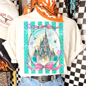 May include: Off-white t-shirt with a fairytale castle illustration in a teal oval, decorated with pink bows and stars. The design includes the text "Magical Pixel Prints" and is set against teal stripes and black polka dots.