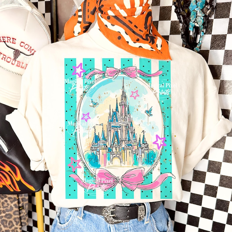 May include: Off-white t-shirt with a fairytale castle illustration in a teal oval, decorated with pink bows and stars. The design includes the text "Magical Pixel Prints" and is set against teal stripes and black polka dots.