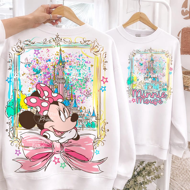 Pode incluir: Duas camisolas brancas com desenhos coloridos da Minnie Mouse. Uma apresenta a Minnie com um grande la&ccedil;o rosa e um castelo, a outra mostra o castelo e as palavras "Minnie Mouse". Ambos os desenhos s&atilde;o emoldurados com bordas douradas e estrelas.