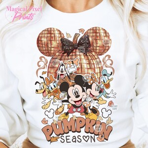 K&ouml;nnte beinhalten: Ein wei&szlig;er Pullover mit einem goldenen Glitzerk&uuml;rbis-Design und einer Gruppe von Disney-Charakteren, darunter Mickey Mouse, Goofy, Donald Duck und Pluto. Der Text "Pumpkin Season" steht unter den Figuren.