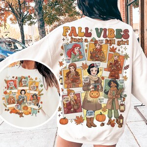 May include: Cream-colored sweatshirt with a fall-themed graphic. The design features the text "FALL VIBES just a fall girl" and illustrations of cartoon princesses in autumn attire. Pumpkins and fall leaves are also included.