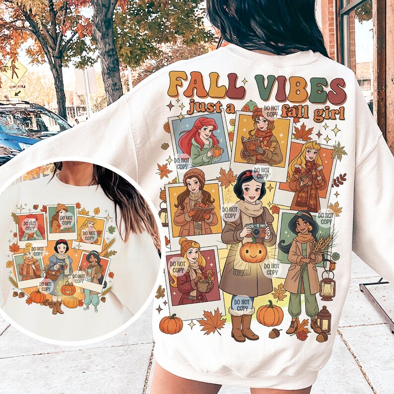 May include: Cream-colored sweatshirt with a fall-themed graphic. The design features the text "FALL VIBES just a fall girl" and illustrations of cartoon princesses in autumn attire. Pumpkins and fall leaves are also included.