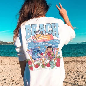Puede incluir: Camiseta blanca de manga larga con un gr&aacute;fico en la espalda. El dise&ntilde;o presenta el texto "BEACH Bum" sobre una ilustraci&oacute;n de Stitch y Lilo en sillas de playa, con olas, una puesta de sol y fresas.