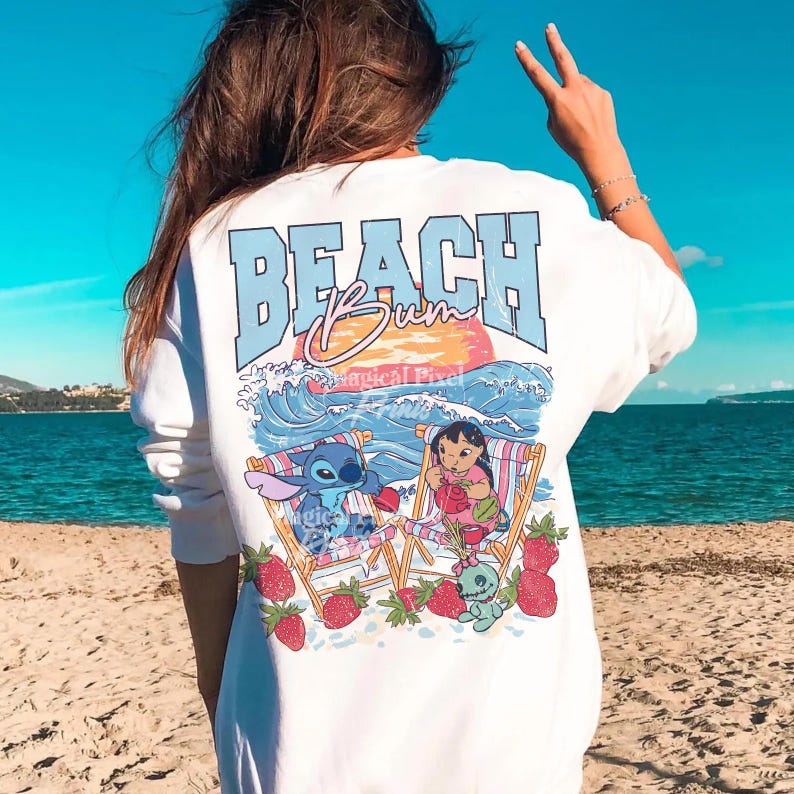 Puede incluir: Camiseta blanca de manga larga con un gr&aacute;fico en la espalda. El dise&ntilde;o presenta el texto "BEACH Bum" sobre una ilustraci&oacute;n de Stitch y Lilo en sillas de playa, con olas, una puesta de sol y fresas.