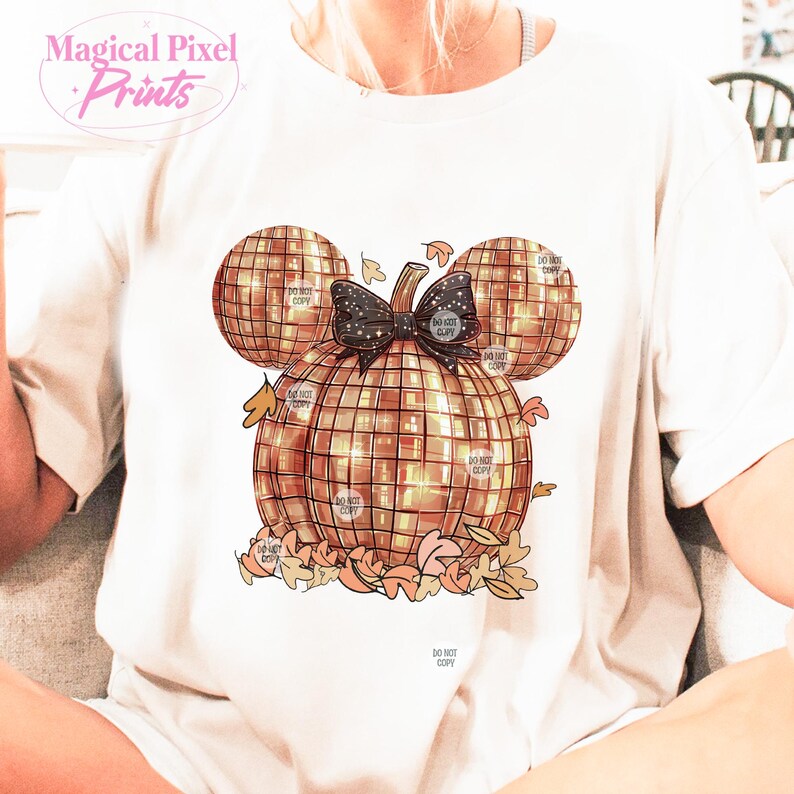K&ouml;nnte beinhalten: Ein wei&szlig;es T-Shirt mit einem goldenen Glitzerk&uuml;rbis-Design. Der K&uuml;rbis ist in Form von Mickey Mouse Ohren und hat eine schwarze Schleife. Das Design ist von Herbstbl&auml;ttern umgeben.