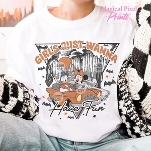 May include: White t-shirt featuring a vintage-style graphic of two cartoon characters driving a car. The text "Girls Just Wanna Have Fun" is printed in orange and black.