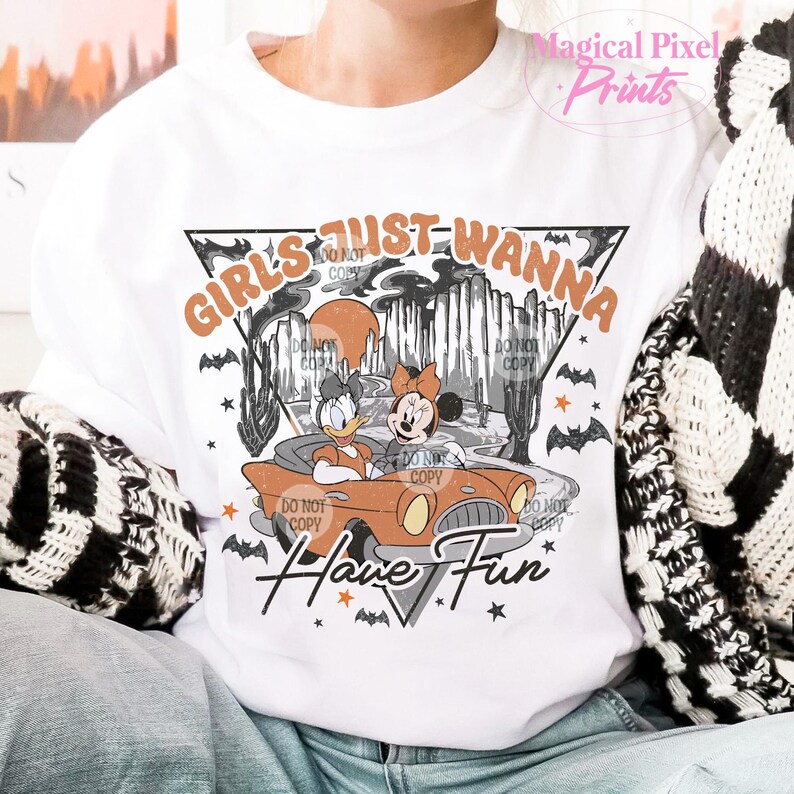 May include: White t-shirt featuring a vintage-style graphic of two cartoon characters driving a car. The text "Girls Just Wanna Have Fun" is printed in orange and black.