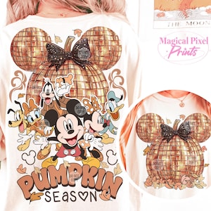 K&ouml;nnte beinhalten: Ein wei&szlig;es T-Shirt mit einem goldenen Glitzer-K&uuml;rbis-Design. Der K&uuml;rbis ist in Form von Mickey-Maus-Ohren und hat eine schwarze Schleife. Das Shirt zeigt auch eine Gruppe von Disney-Charakteren, darunter Mickey Mouse, Minnie Mouse, Donald Duck, Goofy und Pluto. Der Text "Pumpkin Season" ist in orangefarbenen und braunen Buchstaben unter den Figuren geschrieben.