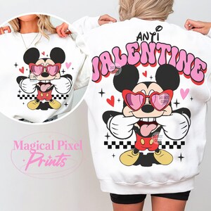 Pu&ograve; includere: Felpa bianca con una grafica rosa "Anti Valentine" e un cartone animato di Topolino che indossa occhiali a forma di cuore. Topolino si sta coprendo la bocca con le mani e sta tirando fuori la lingua.