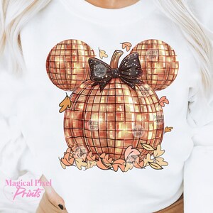 K&ouml;nnte beinhalten: Ein wei&szlig;es Sweatshirt mit einem goldenen Glitzerk&uuml;rbis-Design in Form von Mickey Mouse-Ohren. Der K&uuml;rbis hat eine schwarze Schleife und ist von orangefarbenen und braunen Bl&auml;ttern umgeben.