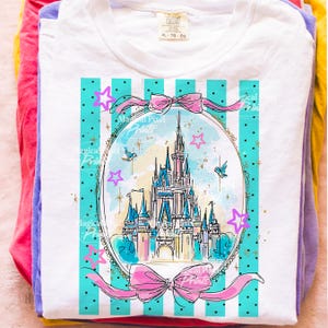 May include: White t-shirt featuring a castle graphic within an oval frame, decorated with pink bows and stars. The design has a blue and white striped background. The shirt is folded on top of other colourful shirts.
