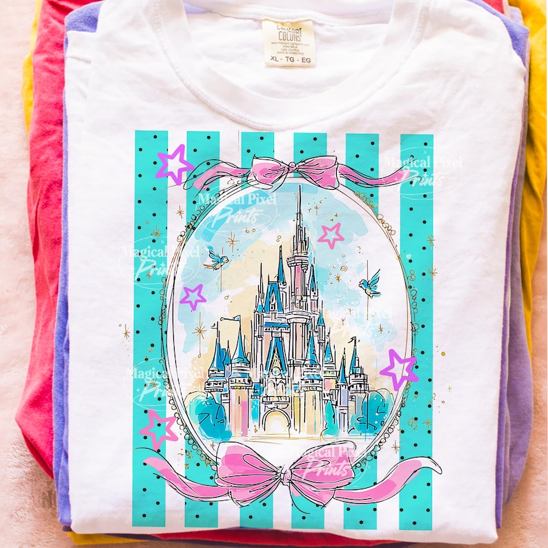 May include: White t-shirt featuring a castle graphic within an oval frame, decorated with pink bows and stars. The design has a blue and white striped background. The shirt is folded on top of other colourful shirts.