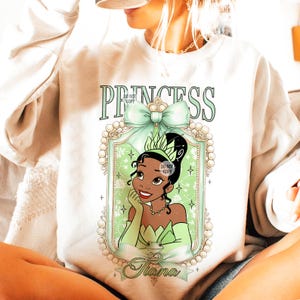 May include: A white sweatshirt with a green and gold princess design. The design features a cartoon image of Princess Tiana from Disney's The Princess and the Frog. The text "Princess" is above the image and the name "Tiana" is below the image.
