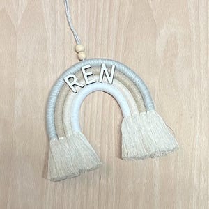 May include: A beige and white macrame rainbow wall hanging with the name "REN" in white letters.