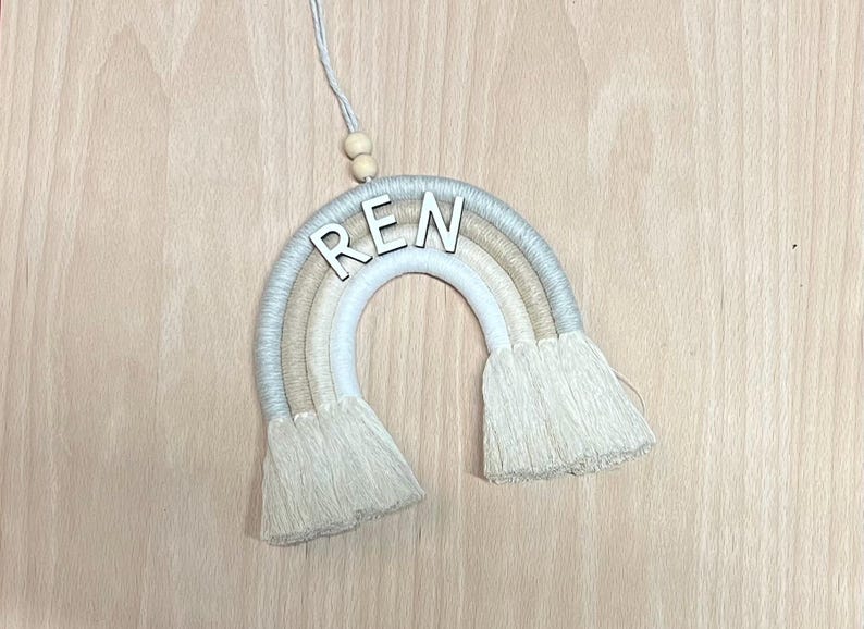 May include: A beige and white macrame rainbow wall hanging with the name "REN" in white letters.