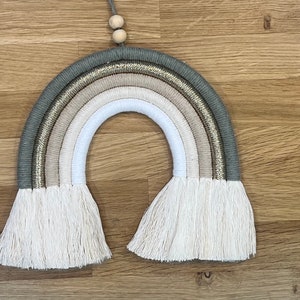 May include: A macrame rainbow wall hanging with a beige, white, and gray color scheme. The rainbow has a gold glitter accent and white fringe.