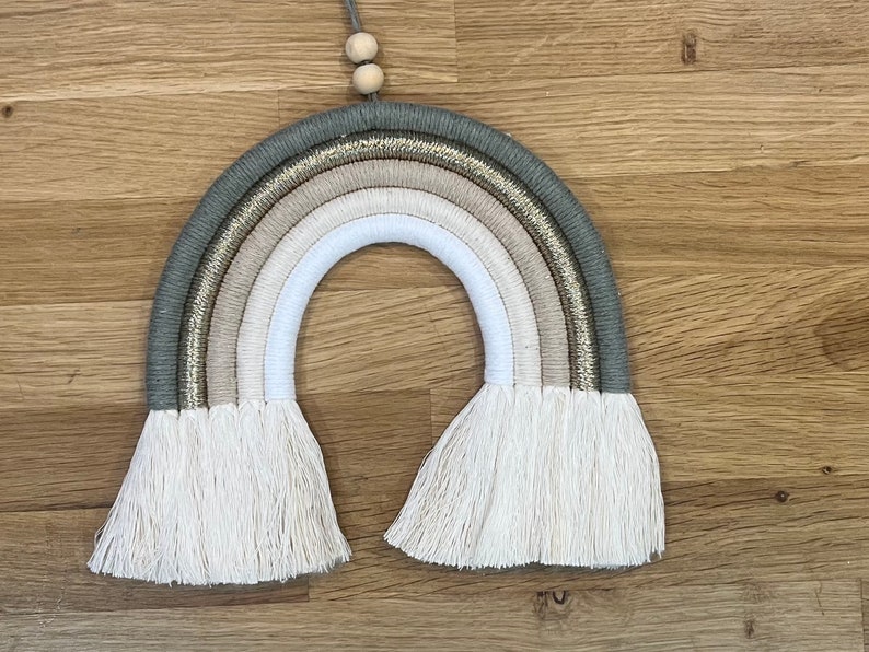 May include: A macrame rainbow wall hanging with a beige, white, and gray color scheme. The rainbow has a gold glitter accent and white fringe.