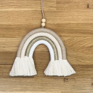 May include: A white, beige, and gold macrame rainbow wall hanging with a wooden bead hanger.
